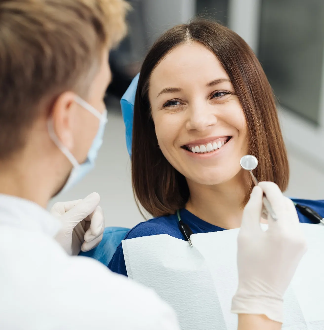 Male professional dentist with gloves and mask performs dental exam on smiling female patient